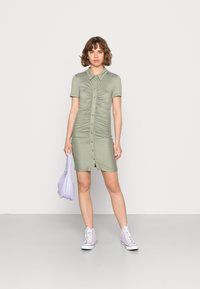 Vestido verde oliva de manga curta com golas, fechamento dianteiro por botões e detalhes franzidos, combinado com uma bolsa lavanda e ténis.