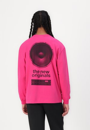 Rosa langermet skjorte med et stort svart høyttalerprint og teksten "de nye originalene" på baksiden. Myk stoff med rund hals.