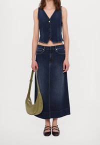 Denimová vesta se třemi zlatými knoflíky, zkombinovaná s dlouhou tmavě modrou denimovou sukní. Doplňky zahrnují zelenou semišovou tašku a tmavé boty.