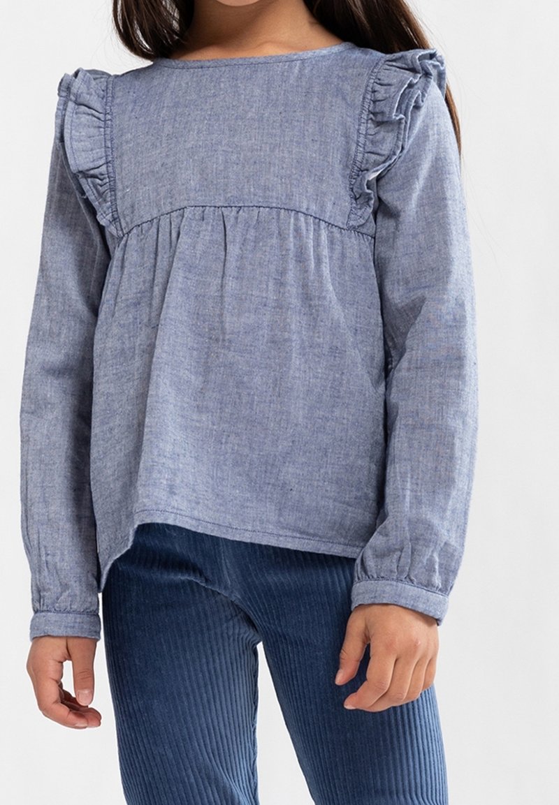 Enfant portant un haut à manches longues bleu avec des volants aux épaules et un pantalon en velours côtelé bleu marine, debout devant un fond blanc.