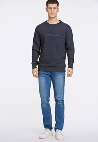 Sudadera de cuello redondo de color marino con el logotipo "LINDBERGH" en el pecho, combinada con jeans celestes y zapatillas blancas. Diseño simple, corte regular, puños acanalados.