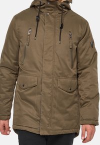 Veste vert olive avec capuche, poches poitrine zippées, fermeture frontale à boutons-pression et deux poches inférieures à rabat. Tissu texturé avec détails renforcés.