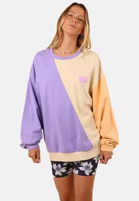 Sweat-shirt coloré, mettant en avant des teintes lavande, crème et pêche, avec une coupe décontractée, des poignets côtelés et un petit patch logo sur le devant.