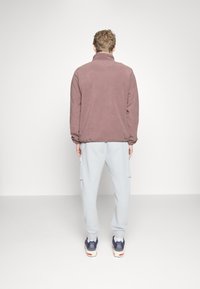 Veste en polaire mauve avec un col haut, associée à un pantalon de survêtement gris clair. La tenue présente une coupe décontractée et une texture.