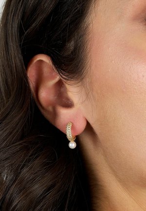 Orecchino a cerchio in tono oro con un intricato design glitterato, caratterizzato da un accento di perla bianca pendente. Superficie texture con finitura lucida.