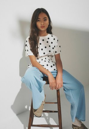 Young woman with long dark hair sitting on a wooden stool, wearing a white polka dot top, light blue wide-leg jeans, and beige shoes.