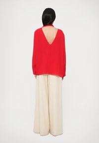 Blouse rouge avec un dos en V profond, associée à un pantalon large couleur crème. La blouse présente des manches longues, un tissu doux et fluide, et un design minimaliste.