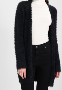 Kvinna som bär en lurvig svart cardigan över en vit ribbad polotröja och högmidjade svarta byxor, står med avslappnade armar.