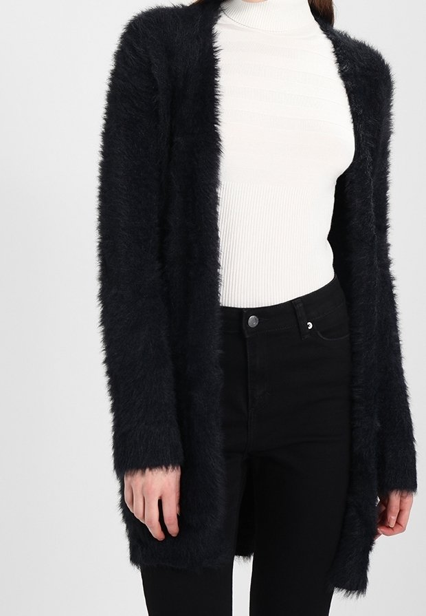 Kvinna som bär en lurvig svart cardigan över en vit ribbad polotröja och högmidjade svarta byxor, står med avslappnade armar.