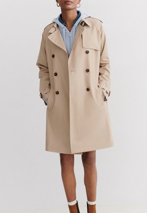 Beige trenchcoat met dubbelzijdige knopen, revers en zijzakken. Draag het over een lichtblauwe hoodie, die een relaxed fit en een gemiddelde lengte heeft.