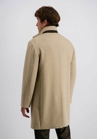 Beige Wollmantel mit hohem Kragen und braunen Akzenten. Gerader Schnitt mit sichtbaren Nähten und glatter Textur. Tragen über karierter Hose.