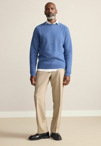 Hellblauer Wollpulli mit Rundhalsausschnitt, zu beigefarbenen Hosen und schwarzen Leder-Schuhen, vor neutralem Hintergrund stehend.