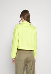 Cropped, bright neon yellow jacket with long sleeves, a pointed collar, and a smooth finish. Worn over khaki pants.