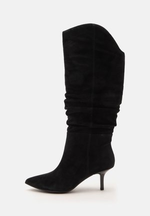 Botte mi-haute en daim noir avec tige souple, bout pointu et talon fin de hauteur moyenne sur fond blanc uni.