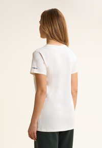 Camiseta blanca de manga corta con cuello redondo y un logo negro en la manga izquierda. Tejido de algodón suave, corte relajado y un brillo sutil.