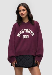 Overmaatse kastanjebruine trui met geribbelde manchetten en onderrand, met witte gestructureerde letters "WRSTBHV" en "030" op de voorkant. Draagt over een gewatteerde zwarte rok.