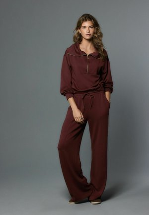 Costum bordo în două piese, format dintr-un top cu glugă și fermoar și pantaloni cu picioare largi. Confecționat dintr-un material moale, cu croială lejeră și buzunare laterale.