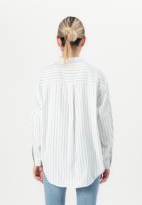 Person med blondt hår i en knute, iført en hvit langermet skjorte med tynne svarte vertikale striper og lyseblå jeans, vendt bort.