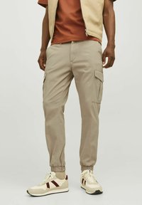 Beige cargo pants with elastic cuffs, multiple side pockets, and a relaxed fit, paired with cream sneakers featuring maroon stripes.