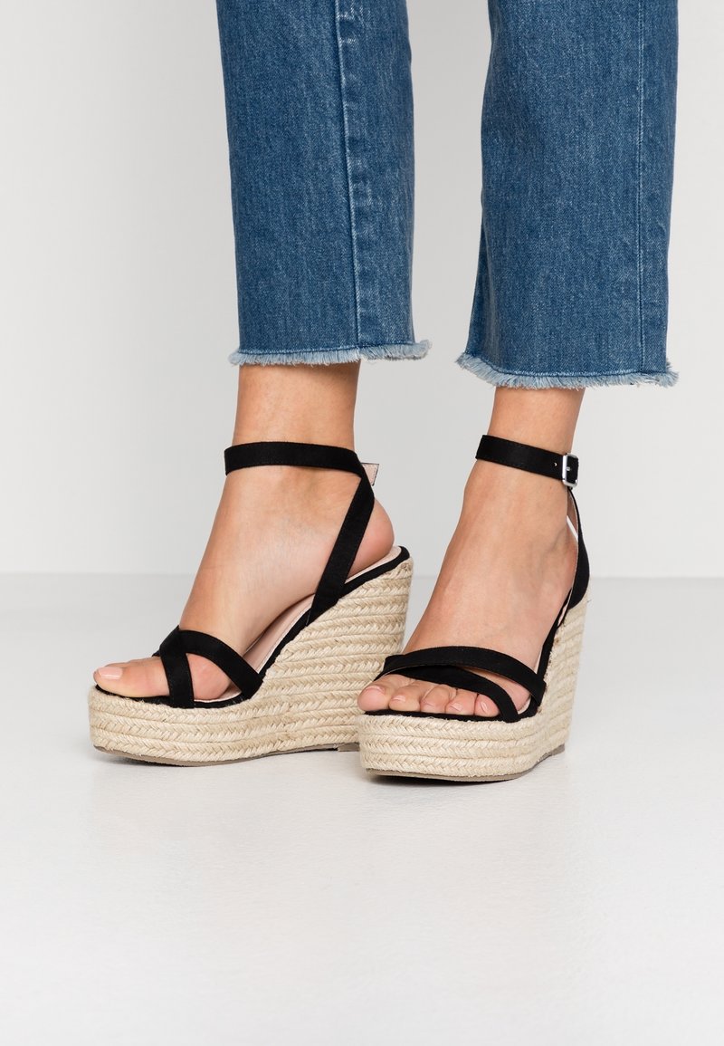 Pieds portant des sandales compensées noires à bride cheville avec semelles en corde tressée, associées à un jean bleu court aux ourlets effilochés.