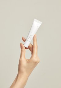 Hvit tube med flip-sekk, merket "NEEDLY." Holdes av en hånd mot en nøytral bakgrunn. Glatt tekstur, minimalistisk design.
