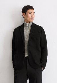 Schwarzer Blazer mit subtiler Textur, mit Reverskragen und zwei Taschen vorne, getragen über einem hellgrauen Sweater mit Reißverschlusskragen. Maßgeschneiderte Passform und klare Linien.