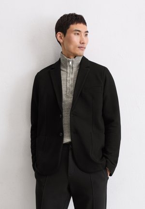 Zwarte blazer met subtiele textuur, notch-lapels en twee voorzakken, gedragen over een lichtgrijze sweater met ritssluiting. Tailored fit en schone lijnen.
