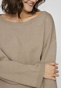 Bež oversize pletena pulover s širokim izrezom, rebrastimi manjšimi rokavi in mehkimi vlakni, prikazan z crossed arms proti svetli ozadju.