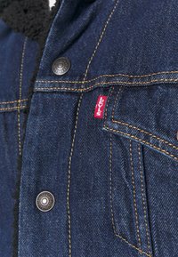 Denim jacket with a dark blue finish, featuring gold stitching, black snap buttons, and a red Levi's label on the front pocket.