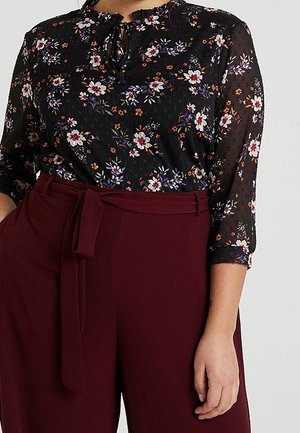 Kvinde iført en sort blomstret bluse med 3/4 ærmer, stoppet ned i højtaljede bordeauxbukser med et matchende stofbælte.