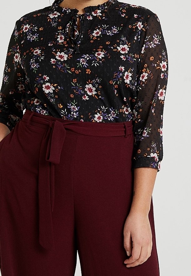Femme portant un chemisier noir à motifs floraux avec des manches trois-quarts, rentrée dans un pantalon taille haute bordeaux avec une ceinture en tissu assortie.