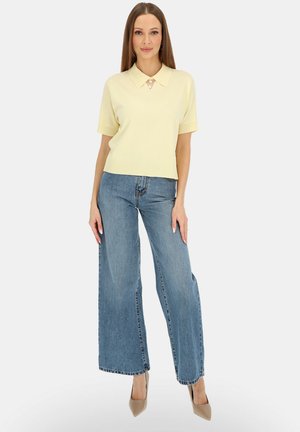 Kobieta ubrana w bladożółty sweter z krótkim rękawem i kołnierzykiem, niebieskie dżinsy o szerokich nogawkach oraz beżowe szpilki w szpic, stojąca na tle białego tła.