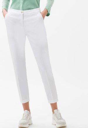 Personne portant un pantalon blanc ajusté aux chevilles avec des poches et des baskets blanches, mains dans les poches, et un haut vert à manches longues visible.