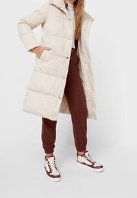 Abrigo acolchado largo color crema con capucha, que presenta un diseño acolchado y botones a presión; llevado sobre joggers marrones y zapatillas marrones y blancas.