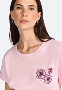 Camiseta de manga corta rosa hecha de tela suave, con un diseño de bordado floral en color púrpura oscuro en la zona del pecho.