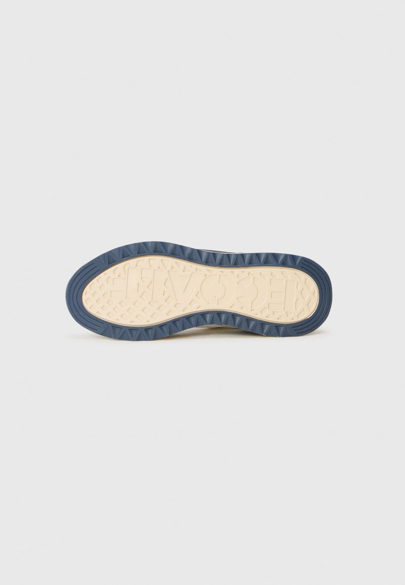 Suela de una zapatilla con un centro beige y bordes azul marino, que presenta patrones geométricos texturizados y letras en relieve del nombre de la marca.