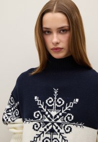 Pull tricoté bleu marine et crème avec un col haut, présentant un motif flocon de neige blanc orné de volutes complexes et de formes géométriques.