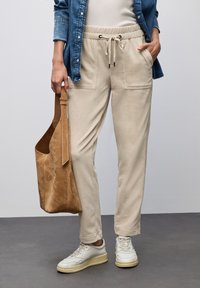 Person i beige dragsko-byxor, vita sneakers, denimskjorta, håller en stor handväska i tan mocka mot en enfärgad bakgrund.