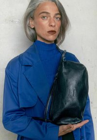 Donna con capelli grigi che indossa un giubbotto blu e un dolcevita tiene una grande borsa in pelle nera con texture contro uno sfondo chiaro.