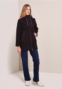 Manteau en polaire violet foncé avec capuche, fermeture éclair à l'avant et deux poches latérales ; associé à un pantalon marine et des baskets blanches.