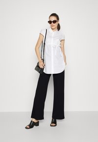White button-up shirt with short sleeves and a tie at the waist, paired with black wide-leg pants. Model wears black open-toe heeled mules and sunglasses.
