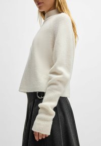 Weißer Strickpullover mit hohem Kragen, gerippten Bündchen und lockerer Passform. Der Stoff erscheint weich und zeigt minimalen Markenschriftzug am Saum.
