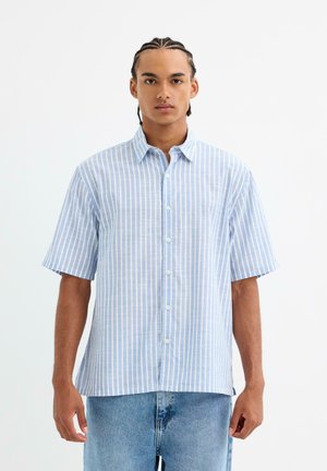 Camisa de manga corta a rayas en azul claro y blanco. Diseño de botones, tela suave, corte relajado. Llevada con pantalones cortos de mezclilla azules.