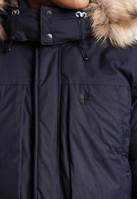 Marinblå isolerad parka-jacka med en huva fodrad med konstfuskpäls, dubbla knappar i fickorna och dragskojusteringar vid kragen.