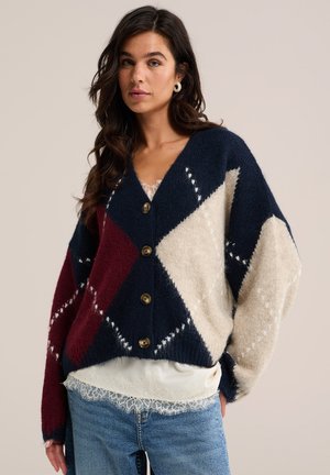 Frau trägt eine blau-burgunderrote-cremefarbene argyle gemusterte Strickjacke mit Knöpfen, mit weißem Spitzenoberteil und blauer Jeans vor schlichtem Hintergrund.