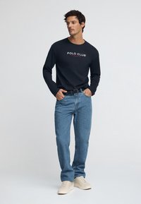 Marinblå tröja med långa ärmar och "POLO CLUB"-logotyp, kombinerad med ljusblå jeans och beige sneakers. Enkel, avslappnad design med klassisk passform.