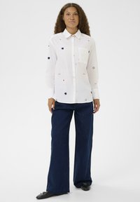 Göndör hajú nő fehér, kis virágmintás gombos ingben és sötét, széles szárú nadrágban, egy sima fehér háttér előtt áll.