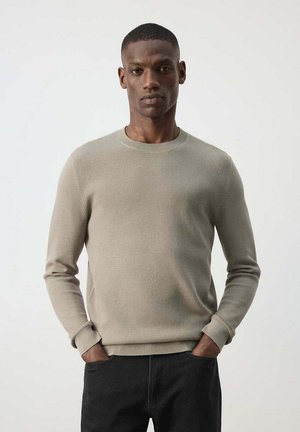 Jonge man met een donkere huid die een lichtbeige textuursweater en een zwarte broek draagt, staand met handen in de zakken tegen een effen achtergrond.