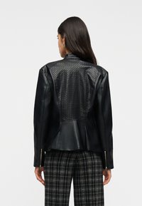 Γυναίκα που φαίνεται από πίσω, φοράει μαύρο δερμάτινο μπουφάν με ραμμένο πίσω πάνελ σε μοτίβο διαμαντιού και καρό παντελόνι.