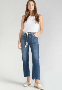 Jean en denim bleu moyen, taille haute et coupe large, avec poches avant et ourlet effiloché, associé à un débardeur en maille blanche.